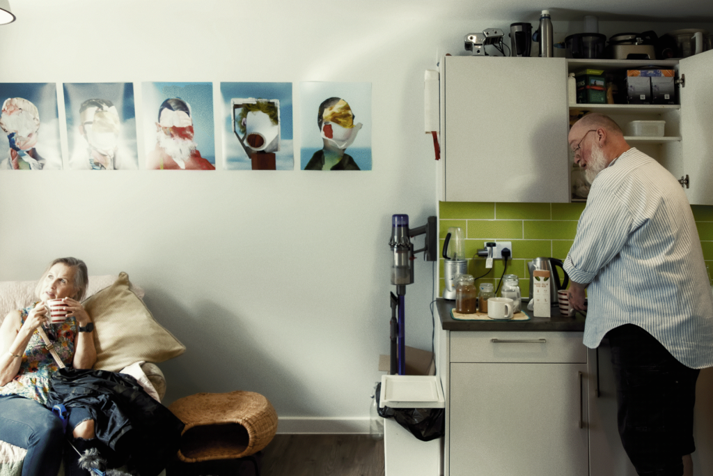 A woman relaxes on a couch while a man prepares coffee in the nearby kitchen. Art hangs on the wall behind them.