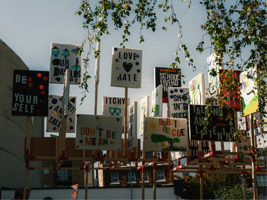 Colorful protest signs with messages promoting love, acceptance, and individuality are displayed outdoors on a kiosk-like structure