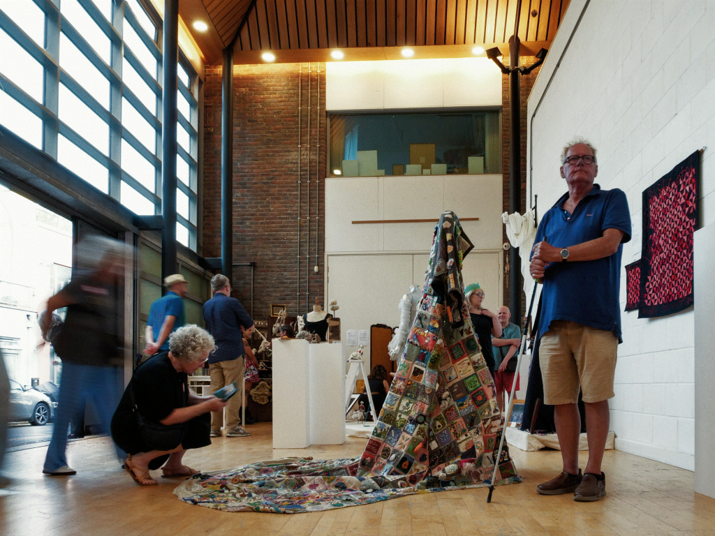 a woman takes a picture of a large quilt on display in a busy gallery