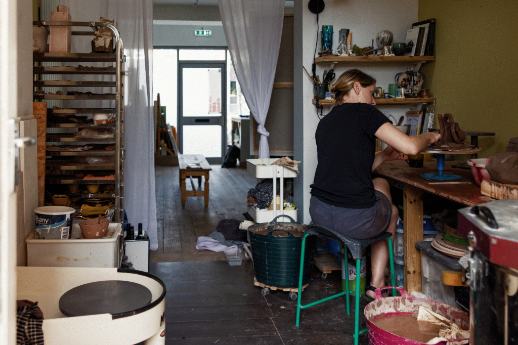 An artist is engaged in creating her work. She is sat at a tall table, around her is clay-related equipment