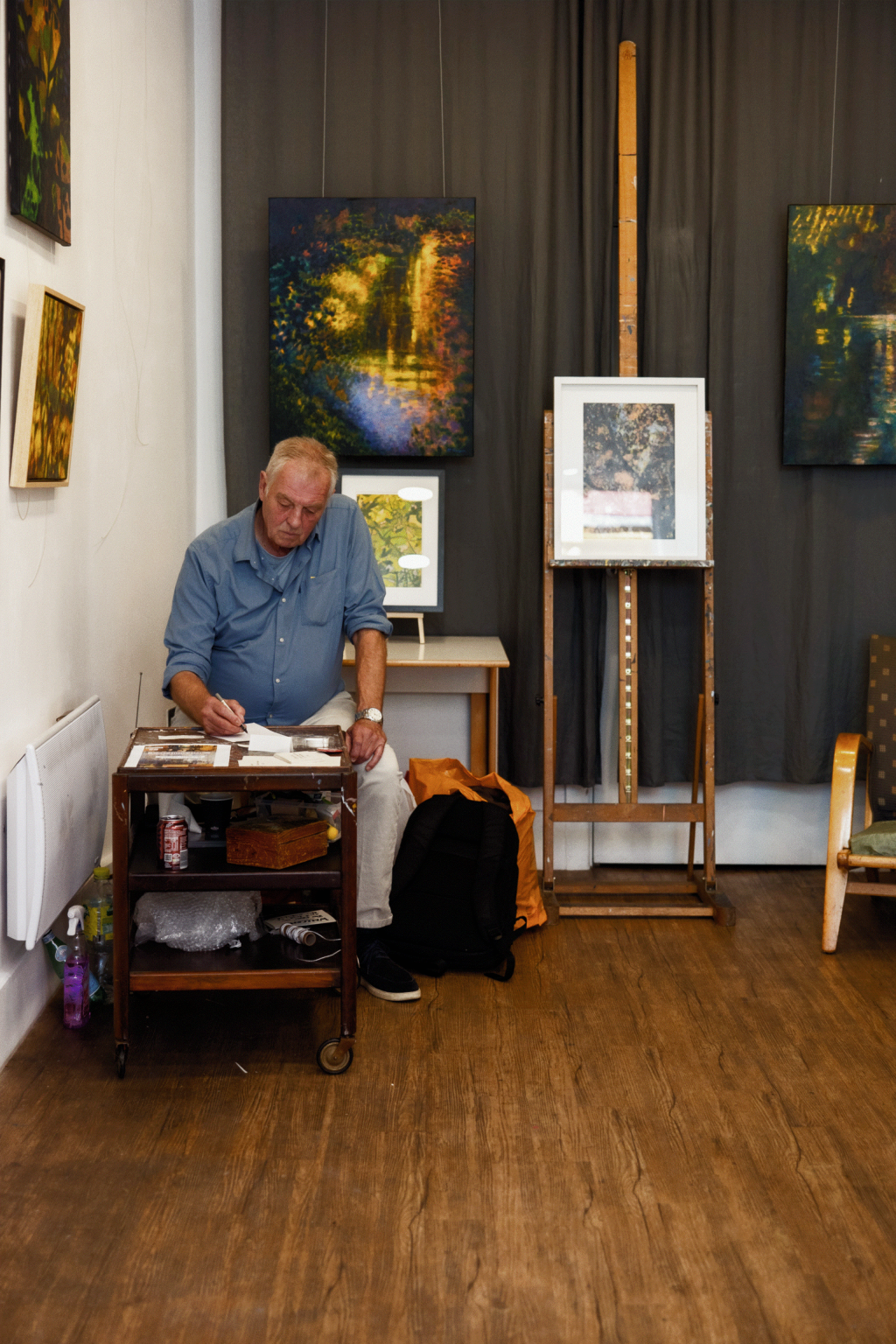 Artist sits at a small table in a studio, sketching while surrounded by his paintings on the walls.