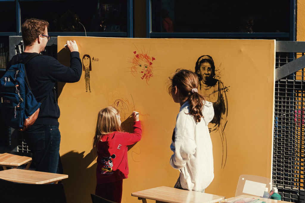 A man and two children are drawing and creating art on a community board