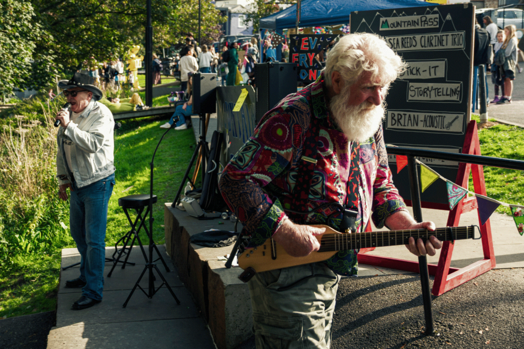 A man plays a string instrument on a stage, while another person stands nearby, singing. The background shows a crowd enjoying a festive outdoor event.