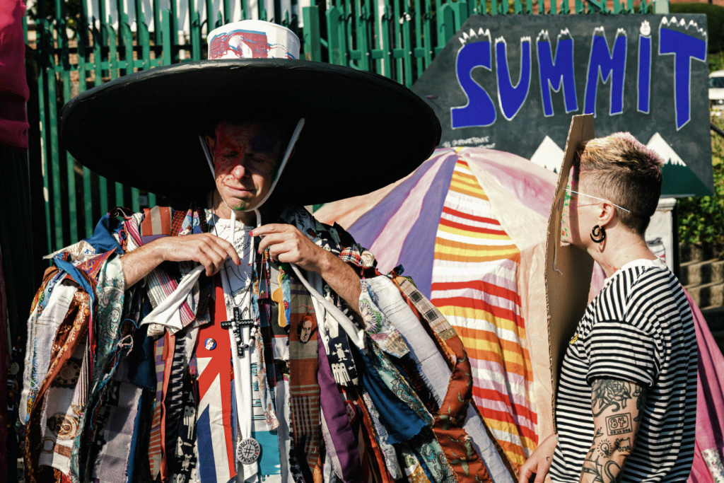 Person dressed in a large hat and a coat made of hundreds of different pieces of colourful fabric, stands next to another person with a cardboard mask on their face